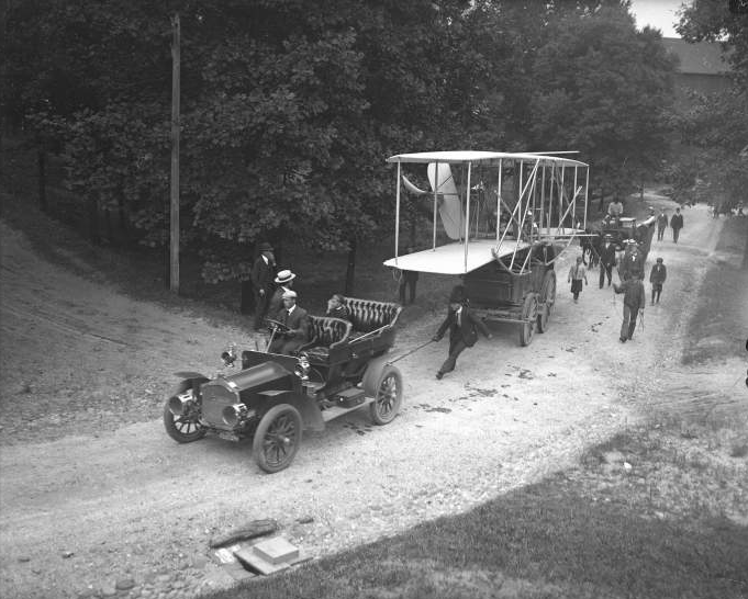 an automobile towed the Wright (Co) Type A Military biplane, with front struts and tail folded for transport, from the storage shed to the Ft. Myer airfield in 1908